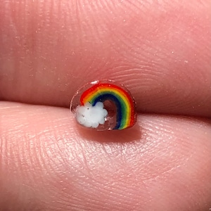Puede incluir: Una pequeña cuenta de arcoíris con una nube blanca. El arcoíris presenta bandas de rojo, naranja, amarillo, verde y azul. La cuenta es transparente y parece estar hecha de vidrio o resina.