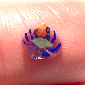 Può includere: Una miniatura di granchio con corpo azzurro, chele blu scuro e dettagli gialli. Il minuscolo granchio è posto su uno sfondo color carne, che mette in risalto i suoi delicati dettagli e la sua maestria artigianale. Il granchio è un oggetto a tema marino.
