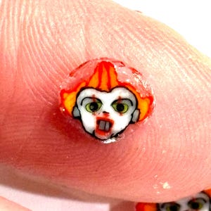 Puede incluir: Una pequeña pegatina redonda roja y amarilla con una cara blanca de payaso con una boca roja y ojos verdes. El payaso sonríe mostrando los dientes.