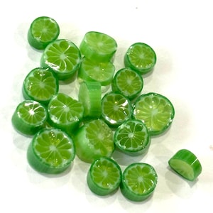 Puede incluir: Una colección de caramelos verdes con forma de lima, cada uno cortado en rodajas. Los caramelos son translúcidos y tienen un acabado brillante.