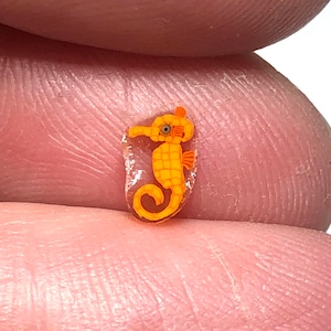 Puede incluir: Una pequeña figura de caballito de mar naranja con un diseño detallado. El caballito de mar tiene un hocico largo, un cuerpo curvado y un ojo pequeño. La figura está dentro de una resina transparente en forma ovalada.