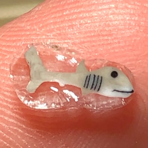 Può includere: Una minuscola statuina a forma di squalo grigio chiaro con strisce e occhi neri. Lo squalo è racchiuso in una sostanza trasparente simile a gel, forse un adesivo o un abbellimento. L'immagine è un primo piano, che mostra i dettagli dello squalo in miniatura.