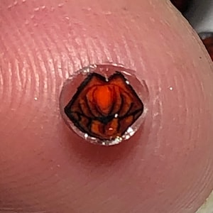 Puede incluir: Una pequeña pegatina redonda y transparente con un diseño rojo y negro en forma de corazón estilizado.