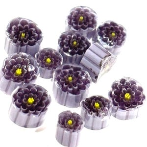 May include: A collection of small, purple glass flower beads with yellow centers. The beads are arranged in a random pattern on a white surface.