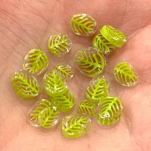 Puede incluir: Una colección de pequeños amuletos de vidrio transparente en forma de hoja con un contorno amarillo verdoso. Los amuletos están esparcidos sobre una mano.