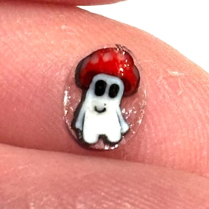 Puede incluir: Una pegatina transparente con un personaje de hongo de dibujos animados. El hongo es rojo con manchas blancas y tiene una cara sonriente blanca con dos ojos negros.