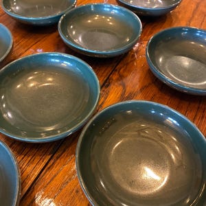 May include: A collection of seven round, dark teal ceramic bowls with a smooth, glossy finish. Each bowl features a simple, shallow design with a slightly raised rim, showcasing a uniform deep green color throughout the set of dinnerware.