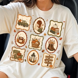 Op de afbeelding: Een off-white t-shirt met een raster van negen herfstachtige illustraties, waaronder een vrachtwagen met pompoenen, een trui, boeken, een kaars, een mand met kalebassen, laarzen, een fauteuil, een lantaarn en een bord met Pumpkin Patch, Apple Cider, Hay Rides.