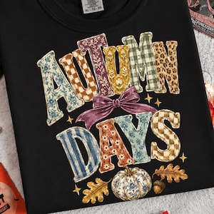 Puede incluir: Una camiseta negra de manga corta con el texto "AUTUMN DAYS" en letras grandes con varios estampados. El diseño incluye un lazo morado, pequeñas estrellas amarillas, hojas de roble y una calabaza con un patrón floral azul y blanco.