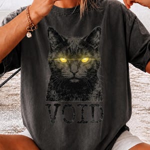 May include: A charcoal gray short-sleeve t-shirt featuring a graphic of a black cat with glowing yellow eyes. The word VOID is printed in large, distressed black capital letters directly below the cat illustration on the front of the shirt.