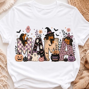 May include: A white short-sleeve t-shirt featuring a graphic print of the word MAMA in large letters. The letters are decorated with dachshunds, ghosts, pumpkins, bats, and flowers in shades of purple, orange, and black. The text Spooky mama is printed on a small black cauldron.
