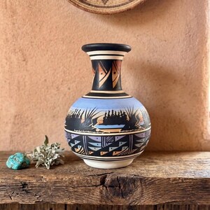 May include: A ceramic vase with a rounded body and a flared neck, featuring a hand-painted landscape of dark silhouettes against a light blue sky, accented by geometric patterns in black, white, tan, and purple around the base and neck.