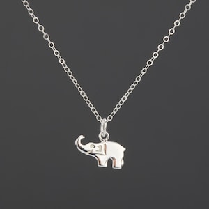 May include: A silver-toned necklace featuring a small, flat elephant-shaped pendant suspended from a delicate cable chain. The elephant charm is oriented to the left with a raised trunk and a smooth, polished metallic finish throughout the piece.