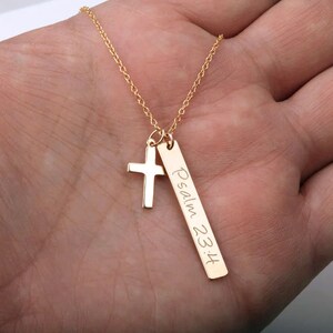 May include: Gold necklace with a small cross charm and a longer bar charm with the inscription "Psalm 23:4".