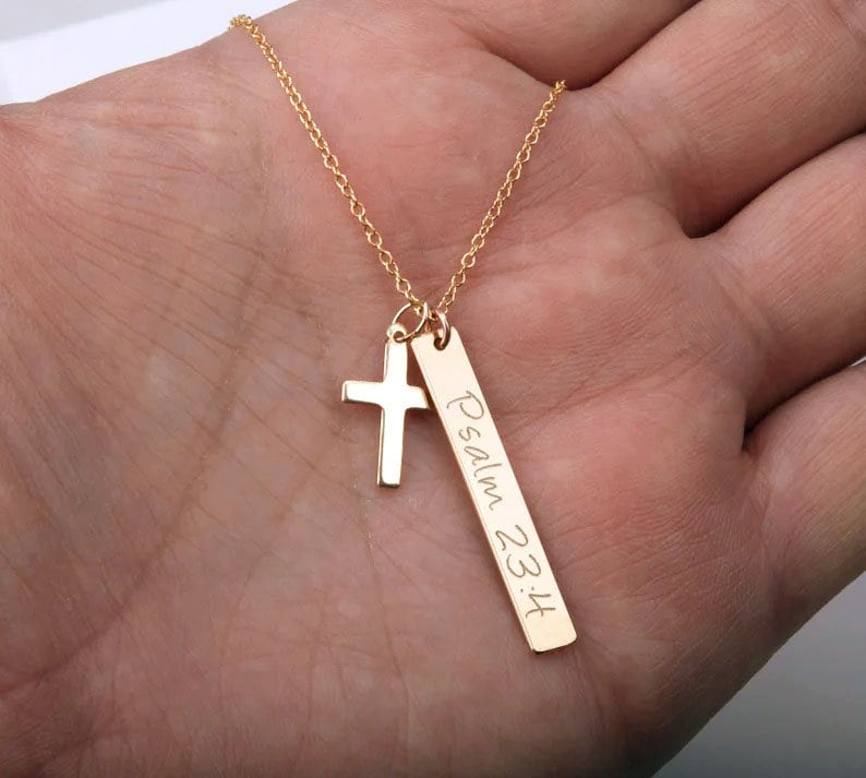 May include: Gold necklace with a small cross charm and a longer bar charm with the inscription "Psalm 23:4".