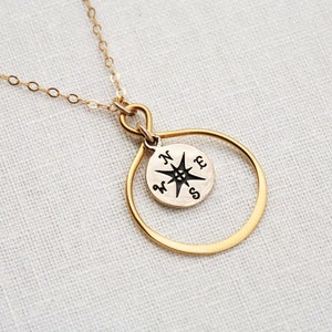 May include: Gold necklace featuring a compass charm and a large, open circle pendant. The compass charm has a black star and the letters N, S, E, and W.