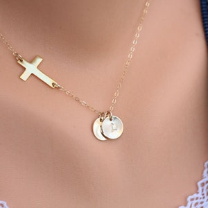 May include: A gold necklace with a delicate cross pendant and two small round charms. The charms have a subtle engraved letter.
