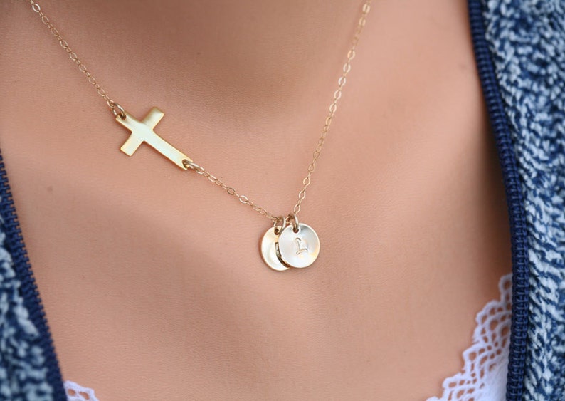 May include: A gold necklace with a delicate cross pendant and two small round charms. The charms have a subtle engraved letter.