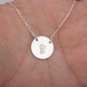 Puede incluir: Un collar de cadena de plata con un colgante redondo que presenta la letra 'C' grabada en una fuente cursiva.
