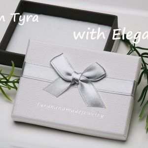 May include: A white gift box with a silver ribbon and bow. The box has the text "Tyrahandmadejewelry" printed on the front. The text "From Tyra with Elegance" is written on the top of the box.