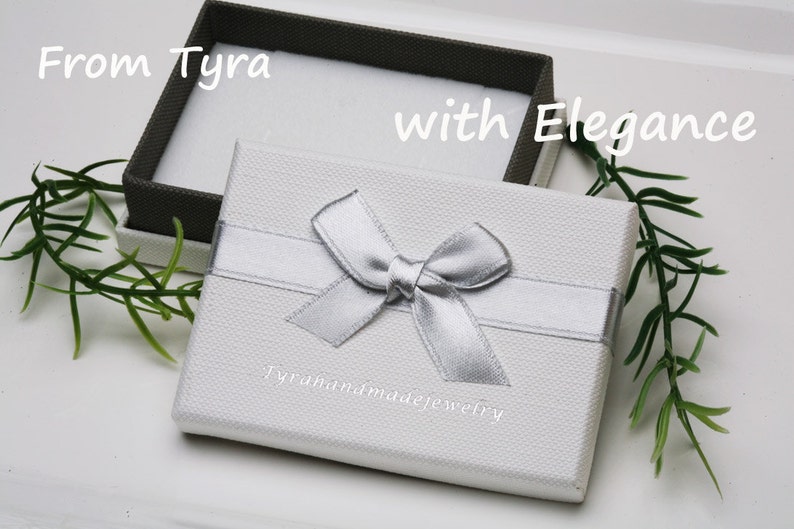 May include: A white gift box with a silver ribbon and bow. The box has the text "Tyrahandmadejewelry" printed on the front. The text "From Tyra with Elegance" is written on the top of the box.
