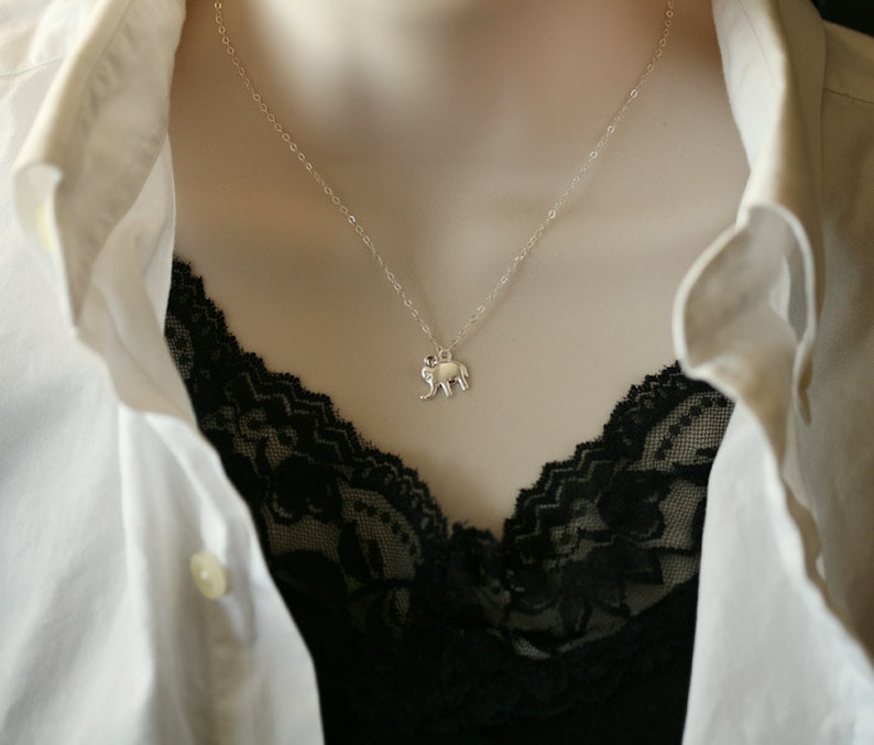 May include: A silver-toned necklace featuring a small, flat elephant pendant suspended from a delicate, thin chain. The pendant is shaped like a stylized elephant in profile, showcasing a smooth, reflective metallic surface and simple, clean lines.