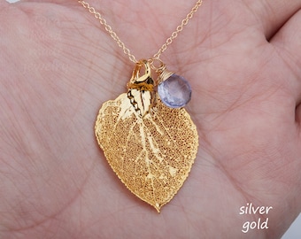 Puede incluir: Collar con colgante de hoja dorada con una cadena delicada. La hoja tiene intrincados detalles de venas y un pequeño dije de piedra preciosa de color púrpura claro. El collar se sostiene en una mano, mostrando el diseño de la joya.