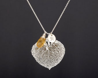 Puede incluir: Un collar de plata con un delicado colgante de hoja de álamo temblón, una pequeña piedra preciosa amarilla y un dije de plata con la letra "M" grabada.