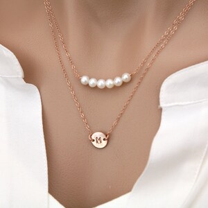 Puede incluir: Un delicado collar de oro con un pequeño colgante redondo con la letra "M" y una fila de cinco perlas blancas.