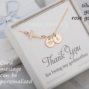 May include: A gold necklace with a cross pendant, a crown charm, and a letter charm. The necklace is in a white box with a thank you message for a godmother. The box is on a white surface.