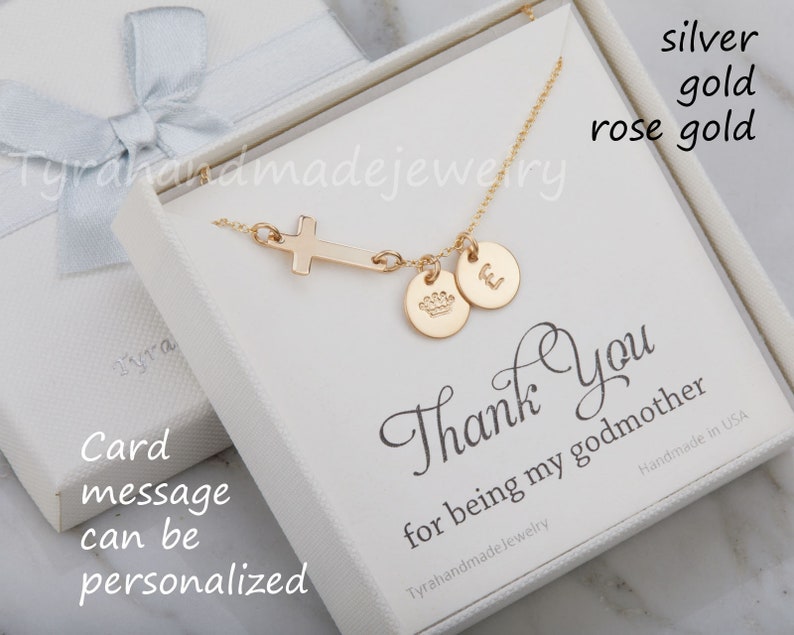 May include: A gold necklace with a cross pendant, a crown charm, and a letter charm. The necklace is in a white box with a thank you message for a godmother. The box is on a white surface.