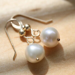Puede incluir: Un par de pendientes de oro con perlas blancas. Los pendientes tienen un diseño sencillo con una pequeña bola de oro en la parte superior de cada pendiente.