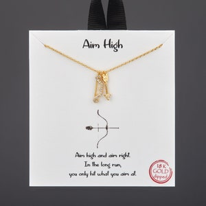 May include: Gold necklace with a bow and arrow charm. The necklace is on a white card with the text "Aim High" and "Aim high and aim right. In the long run, you only hit what you aim at." The card also has a red circle with the text "18K GOLD dipped".