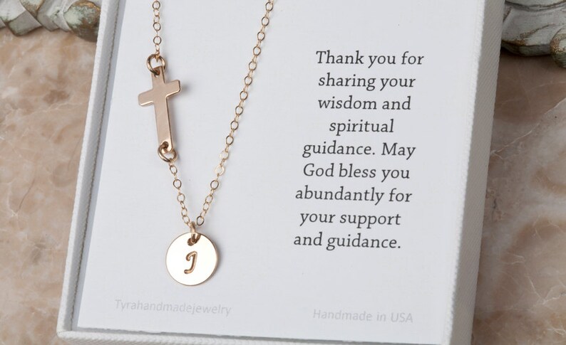 May include: Gold necklace with a small cross charm and a round charm with the letter 'J' engraved on it. The necklace is in a white box with a thank you message.