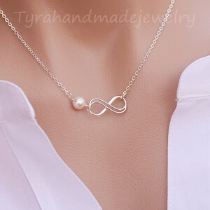 May include: A silver infinity necklace with a single pearl pendant. The necklace is on a white background.
