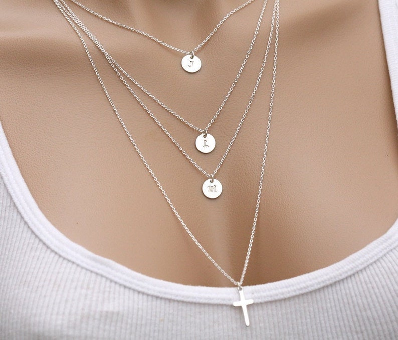 May include: A silver necklace with four pendants. The top pendant is a circle with the letter "J" engraved on it. The second pendant is a circle with the letter "L" engraved on it. The third pendant is a circle with the letters "M" and "L" engraved on it. The bottom pendant is a cross.