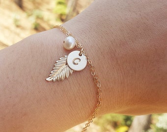 May include: A gold chain bracelet with a delicate feather charm, a pearl bead, and a small round charm with the letter "C" engraved on it.