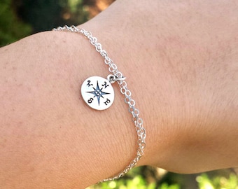 Puede incluir: Un brazalete de cadena de plata con un dije de brújula. El dije de la brújula tiene una estrella de plata en el centro y las letras N, S, E y W alrededor del borde.