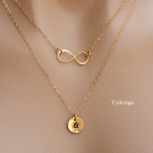 Puede incluir: Collar de oro de dos capas con un dije de símbolo de infinito y un pequeño dije redondo con la letra G. El collar está en un maniquí.
