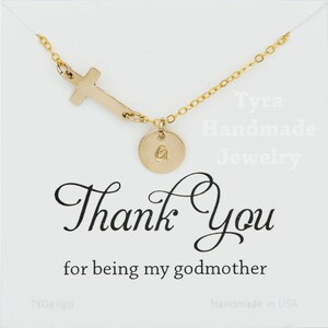 May include: Gold necklace with a cross charm and a small round charm with the letter "G" engraved on it. The necklace is displayed on a white card with the text "Tyra Handmade Jewelry" and "Thank You for being my godmother".