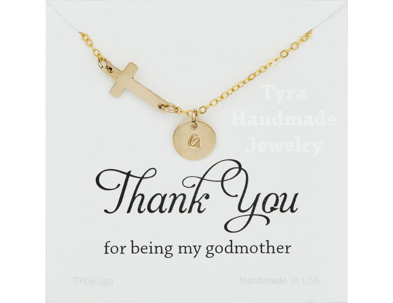 May include: Gold necklace with a cross charm and a small round charm with the letter "G" engraved on it. The necklace is displayed on a white card with the text "Tyra Handmade Jewelry" and "Thank You for being my godmother".