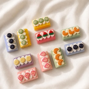 May include: A collection of ten rectangular miniature cake charms in various pastel colours, each topped with white cream dollops and small fruit slices including blueberries, kiwi, strawberries, watermelon, and orange segments.
