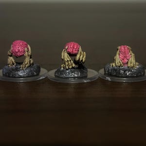 May include: Three miniature figurines of creatures with exposed pink brains and tan, clawed hands. Each figure is mounted on a dark, textured, rocky base attached to a clear, circular plastic disc. The figures are positioned in a row, displaying different angles of the same creature design.