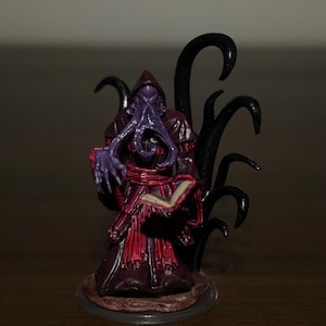 May include: A painted miniature figurine of a hooded creature with a purple tentacled face, wearing a dark red and purple robe. The figure holds an open book in one hand and is surrounded by black, curved, tentacle-like structures behind it.