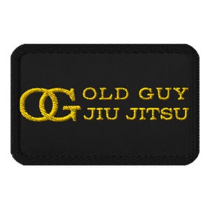 May include: A rectangular black embroidered patch featuring the yellow text OLD GUY JIU JITSU and a large yellow monogram logo consisting of the letters O and G intertwined on the left side, all finished with a thick black stitched border.
