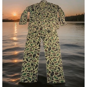 May include: A long-sleeve button-up jacket and matching trousers set featuring a camouflage pattern in shades of olive green, dark brown, and tan. The jacket includes a collared neckline and two front pockets, while the trousers have a straight-leg cut.