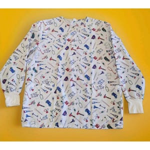 May include: A white long-sleeve button-down scrub top featuring a repeating pattern of medical equipment including stethoscopes, crutches, bandages, scissors, and ambulances in red, blue, and black. The garment has two front pockets and white cuffs.