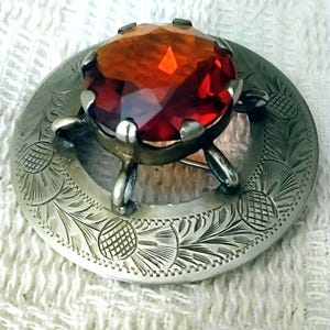 May include: A circular silver-toned brooch featuring an engraved thistle pattern along the outer rim. A large, faceted, deep orange-red gemstone is set in the center, held securely by six metal prongs that rise from the base of the brooch.