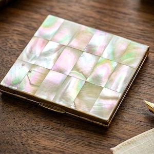 May include: A square compact case featuring a grid of iridescent mother-of-pearl tiles with a gold-toned metal frame. The surface displays shifting hues of pink, green, and white, reflecting light across the rectangular inlay segments.