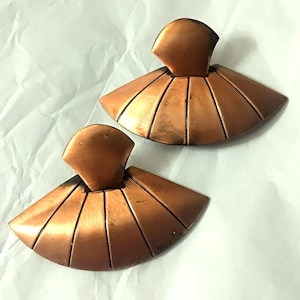 Puede incluir: Un par de pendientes de metal en tono cobre con un diseño en forma de abanico y líneas verticales grabadas. Cada pendiente tiene una sección superior redondeada conectada a una base semicircular más ancha y simétrica.